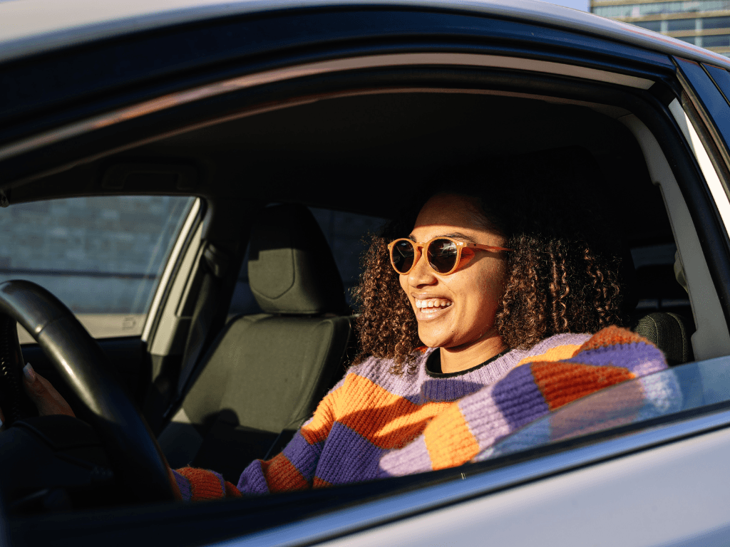 A woman wearing sunglasses driving her car in bright sunlight, illustrating the practical benefits of exploring various types of car cover.